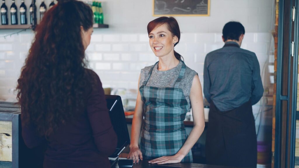 A barista with a plaid apron smiles while interacting with a customer at a café counter.